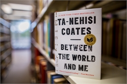 [ai] A book titled "Between the World and Me" by Ta-Nehisi Coates displayed on a shelf in a library. The cover features large black text on a white background with a circular award seal.